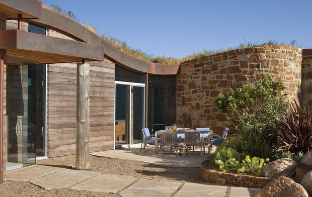 Exterior courtyard view of a low, organically curved contemporary residence. The building combines weathered timber cladding, substantial raw concrete elements, dark-framed glazing, and a curved rough stone wall. A planted roof and native-style landscaping surround an outdoor dining area paved with large irregular stone slabs.