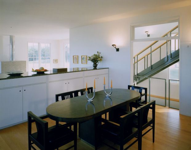 Bright dining area with a large dark oval dining table and matching black ladder-back chairs, positioned in front of white built-in cabinetry. The room has pale walls, timber flooring, framed artwork, and a glazed opening revealing an internal staircase with dark steel stringers and pale wood handrails. Natural light enters through tall windows, creating a clean, refined interior.