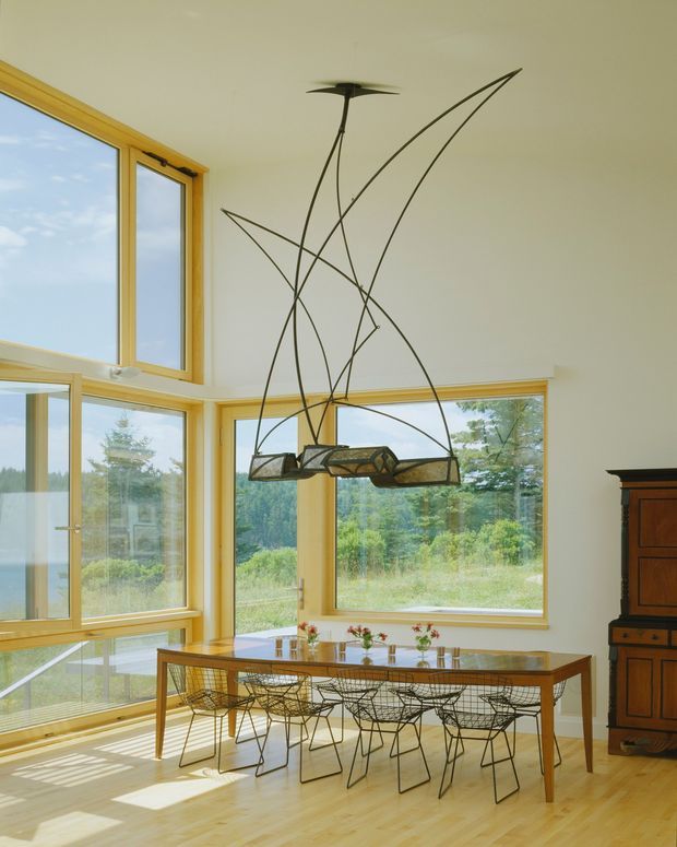 Bright dining area with expansive timber-framed windows overlooking a natural landscape. A long wooden dining table is paired with wire-frame dining chairs, while a sculptural suspended pendant light forms a dramatic focal point above. Pale timber flooring, light walls, and warm wood window frames create an airy, mid-century-inspired interior.