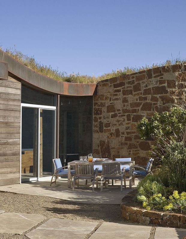 A sunlit outdoor dining terrace enclosed by a curved weathered metal roof edge and a rustic stone wall. Timber-clad glazing and a glass door open onto the paved terrace, which contains a wooden dining table and chairs. Gravel paths, large rectangular paving slabs, and planted garden beds surround the space, with a low planted green roof visible above the structure.