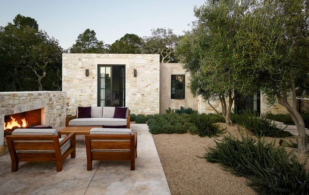 A low-profile stone-clad residence with a landscaped courtyard terrace, outdoor fireplace, and timber lounge furniture. Pale masonry walls, dark-framed glazing, gravel planting beds, olive-like trees, and drought-tolerant shrubs create a warm Mediterranean-style outdoor living setting.