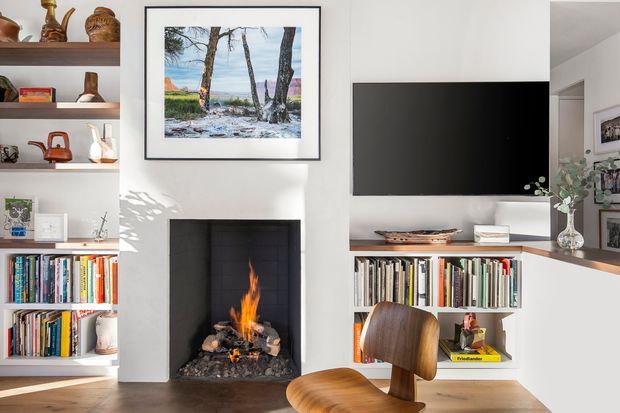 Bright, carefully built-in living room with white shelving and cabinetry framing a central dark fireplace, a wall-mounted television, extensive book storage, and a compact timber desk. A framed landscape artwork, ceramics, plants, and books add warmth to the otherwise clean white interior. The room has timber flooring and a warm wood-and-white palette.