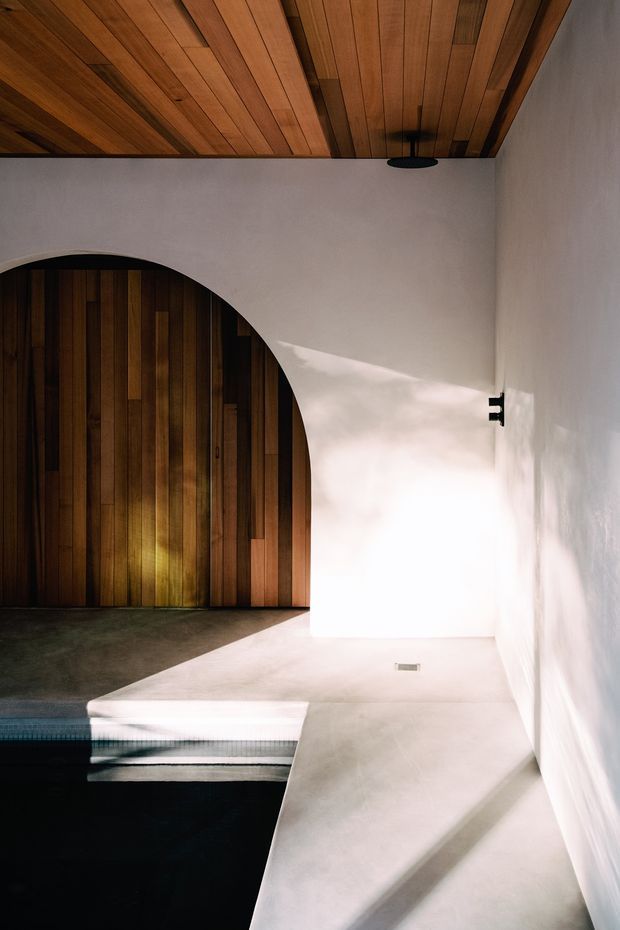 A minimalist architectural interior featuring a dark swimming pool in the foreground, pale concrete or plaster platforms and steps, white rendered walls, and a dramatic arched timber door or wall opening. The ceiling is lined with warm timber boards, creating a strong contrast with the light surfaces. Natural light casts angular shadows across the walls and floor.