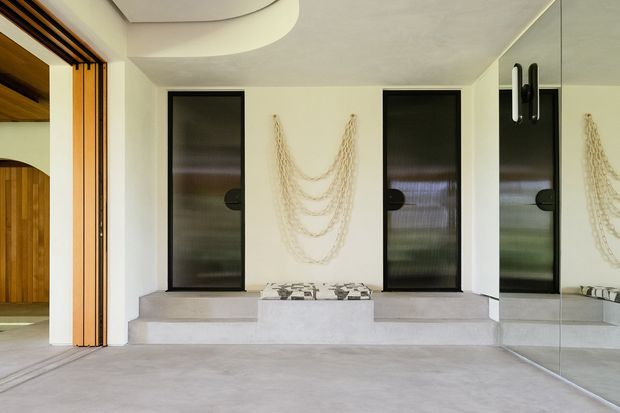 A contemporary entryway or hall with pale smooth flooring and walls, three tall dark-framed glazed doors or panels set into a stepped platform, and a large mirrored wall on the right. A curved recessed ceiling feature and warm timber slatted screening add architectural interest, while delicate hanging rope or chain decor forms a sculptural wall accent.