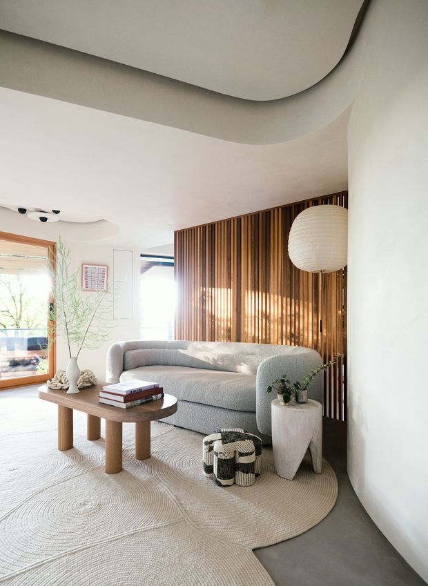 Bright, refined living room with a curved light-grey sofa, rounded timber coffee table, textured neutral rug, sculptural side table, woven pouf, and a large paper pendant light. A vertical timber slatted screen forms a feature wall behind the sofa, while broad curved ceiling details and warm timber-framed glazing add architectural interest.
