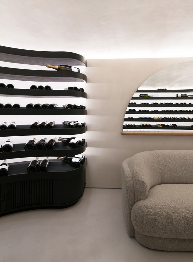 A refined wine room or wine retail display with curved black wall-mounted and freestanding bottle racks, softly lit cream plaster walls, and a sculptural arched mirror or glazed display niche. A rounded beige upholstered lounge chair occupies the foreground, creating a quiet, luxurious hospitality atmosphere.