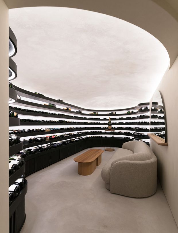 A luxurious, compact eyewear retail showroom with curved, tiered black display shelving wrapping around the walls and filled with glasses. The space features a large sculptural oval/organic-shaped illuminated ceiling, softly curved cream walls, pale flooring, a rounded textured cream sofa, and a small light timber coffee table. The design is highly refined, monochromatic, and softly lit with strong curved forms.