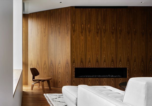 A refined living room featuring a dramatic full-height dark timber veneer feature wall with a built-in linear fireplace. A white sectional sofa occupies the foreground, accompanied by a patterned rug, dark side table, and sculptural timber lounge chair. The room has a clean white ceiling with recessed downlights and a warm timber floor, creating a sophisticated, contemporary interior.