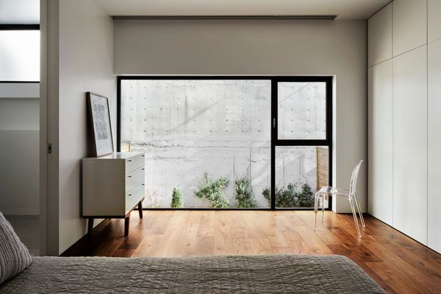 A minimalist bedroom with warm timber flooring, pale built-in wall cabinetry, a low-profile recessed ceiling light, and a large black-framed window. The bed occupies the foreground, while a small mid-century-style cabinet, framed artwork, and two light chairs sit near the glazing. A concrete boundary wall and planting are visible outside.