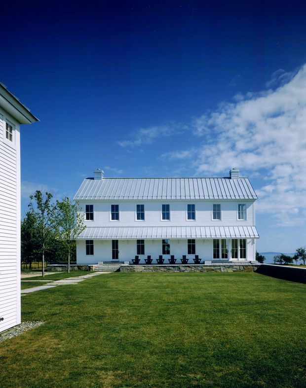 Exterior view of a large, symmetrical white two-storey building with a long pitched metal roof, multiple chimneys, evenly spaced windows, and a continuous covered veranda. The building sits within a broad manicured lawn, with another matching structure partially visible at left and a distant body of water or horizon at right. Clear blue sky and simple white detailing create a restrained coastal or traditional institutional character.