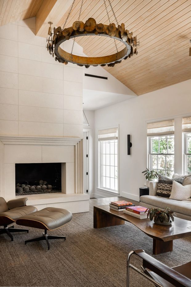 A refined living room with a large white tiled fireplace, exposed light timber vaulted ceiling, tall divided-light windows with woven shades, and a contemporary seating arrangement. A sculptural circular metal-and-wood chandelier hangs prominently overhead, while a leather lounge chair, sofa, substantial timber coffee table, and neutral textured rug create a warm, sophisticated interior.