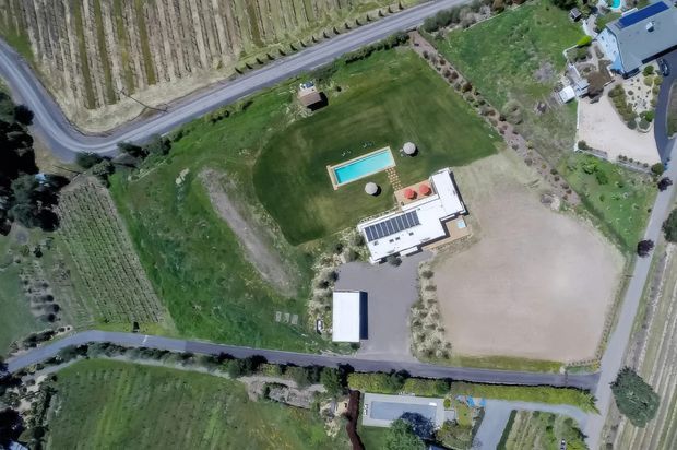 Aerial view of a rural property set among cultivated fields and orchards. The site includes a large modern house with a flat or low-profile roof, a rectangular swimming pool, extensive lawns, landscaped planting, paved driveways, and an outbuilding. Surrounding agricultural land and access roads are clearly visible.