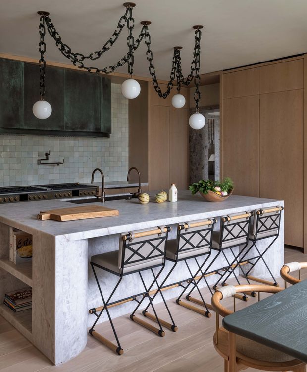 A sophisticated kitchen centered on a large pale stone island with integrated double sinks and dark metal-and-timber bar stools. The room features warm wood floor-to-ceiling cabinetry, a dark aged-metal rangehood, pale square tiled splashback, brass-toned fixtures, and distinctive chain-hung globe pendant lights. A dining table and chairs are partially visible in the foreground.