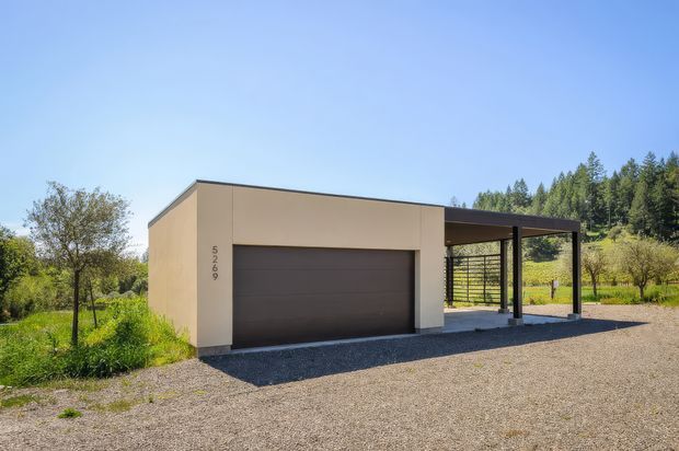 A detached contemporary garage and covered carport structure in a landscaped rural setting. The building has a simple flat-roofed, rectilinear form with smooth beige rendered walls, a wide dark sectional garage door, and a slim dark metal-framed carport canopy. Gravel paving surrounds the structure, with trees and a wooded hillside in the background.
