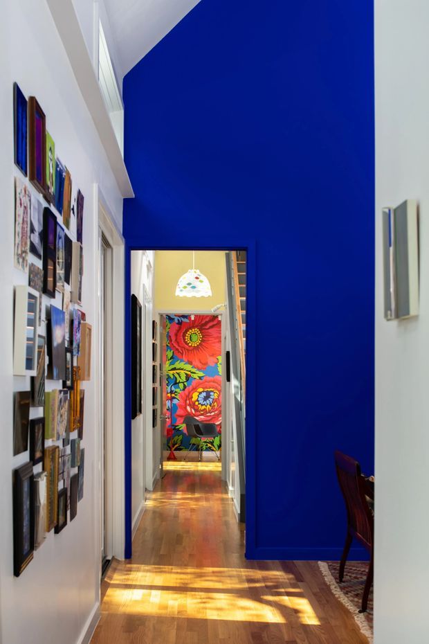 A narrow, double-height residential hallway with vivid cobalt-blue walls, white trim, and warm timber flooring. The left wall is densely arranged with framed artwork and photographs, while a doorway at the far end reveals a brightly coloured floral feature wall and pendant light. Strong sunlight creates patterned highlights across the timber floor, and part of a dark timber chair and patterned rug is visible at right.
