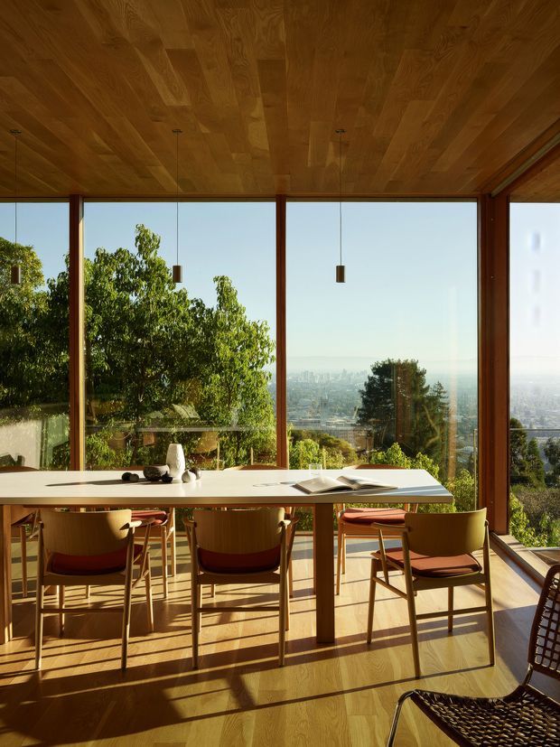A sunlit dining room with a long light-coloured dining table and wooden chairs, enclosed by expansive floor-to-ceiling glazing. The room features warm timber flooring, a timber-clad ceiling, slender dark timber window frames, and simple pendant lights. Trees and a distant cityscape are visible beyond the glass, while strong afternoon light creates long shadows across the floor.