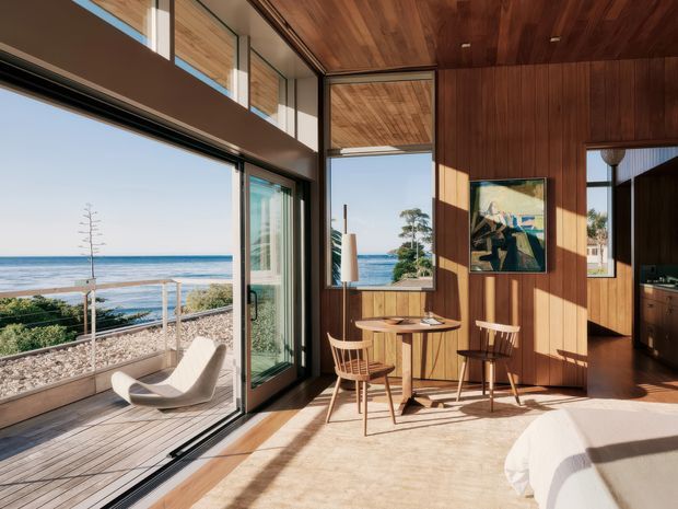 Sunlit coastal living and dining area with full-height glazed sliding doors opening to a timber terrace overlooking the ocean. The room features extensive warm timber wall and ceiling lining, a small round dining table with wooden chairs, sculptural lounge seating, a light rug, and an adjacent kitchen visible through the opening.