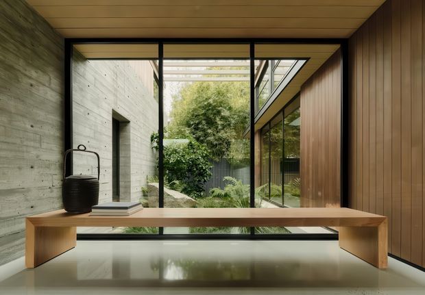 A refined modern interior centered on a long, minimalist timber console or desk in front of full-height black-framed glazing. The space combines a raw concrete wall, warm timber ceiling and vertical timber wall panelling, with a landscaped internal courtyard visible beyond the glass. The restrained palette and clean geometry create a calm, contemporary atmosphere.