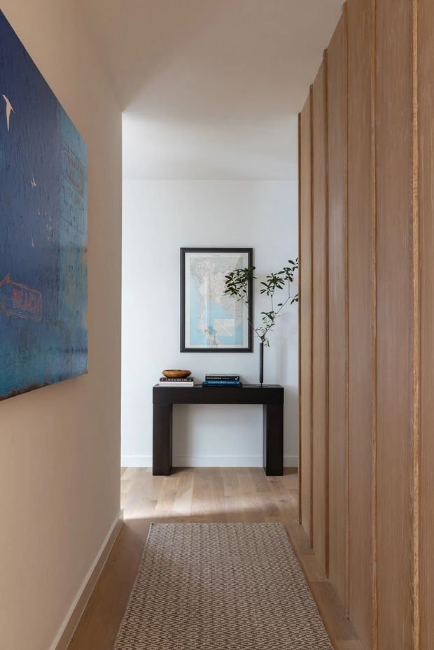 A narrow, softly lit residential hallway or entry passage with pale timber flooring, a large textured blue artwork on the left wall, and full-height vertical timber wall panelling or built-in joinery on the right. At the end is a dark console table styled with books and a slender vase containing greenery, beneath a framed map. A neutral patterned runner extends along the floor, creating a warm, refined and curated entrance.