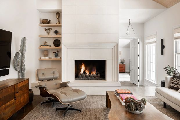 Bright, refined living room centred on a large white tiled fireplace with a lit fire. The room includes a tan leather lounge chair and ottoman, a white sofa, a timber media console, open display shelving, a large timber coffee table, tall windows with blinds, and a partial timber-lined sloped ceiling.
