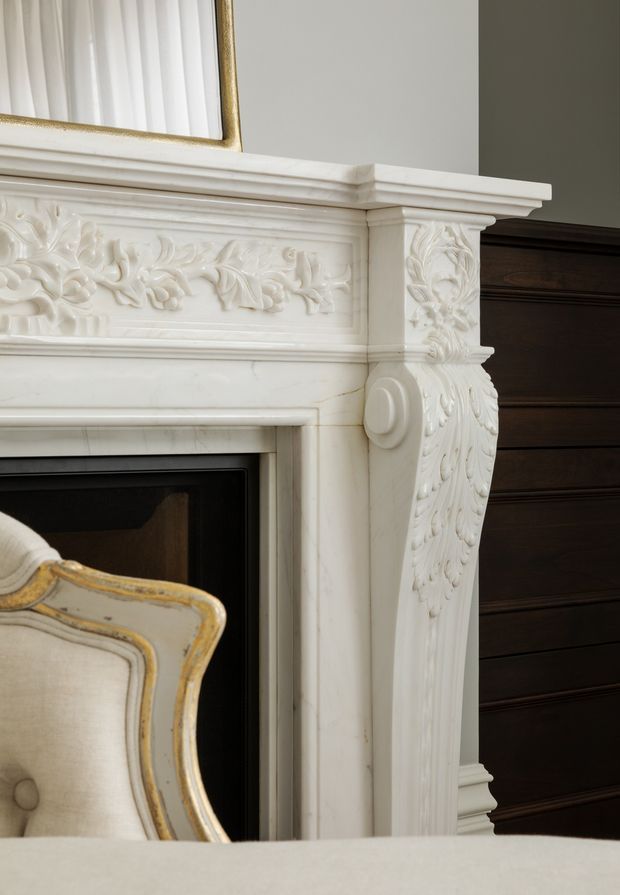 Close-up interior detail of an ornate white carved fireplace mantel with floral reliefs, classical mouldings and column-like side detailing. A gilded framed mirror is partially visible above, with dark timber wall panelling behind and an upholstered chair in the foreground. The image emphasizes refined traditional architectural detailing.