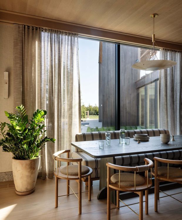 A warm, refined dining area with a built-in upholstered banquette and rounded wooden dining chairs around a rectangular table. Floor-to-ceiling sheer curtains frame large glazing, with a leafy indoor plant, timber ceiling, timber-toned flooring, and a sculptural suspended pendant light. A dark timber-clad exterior wall and courtyard-like view are visible through the glazing.