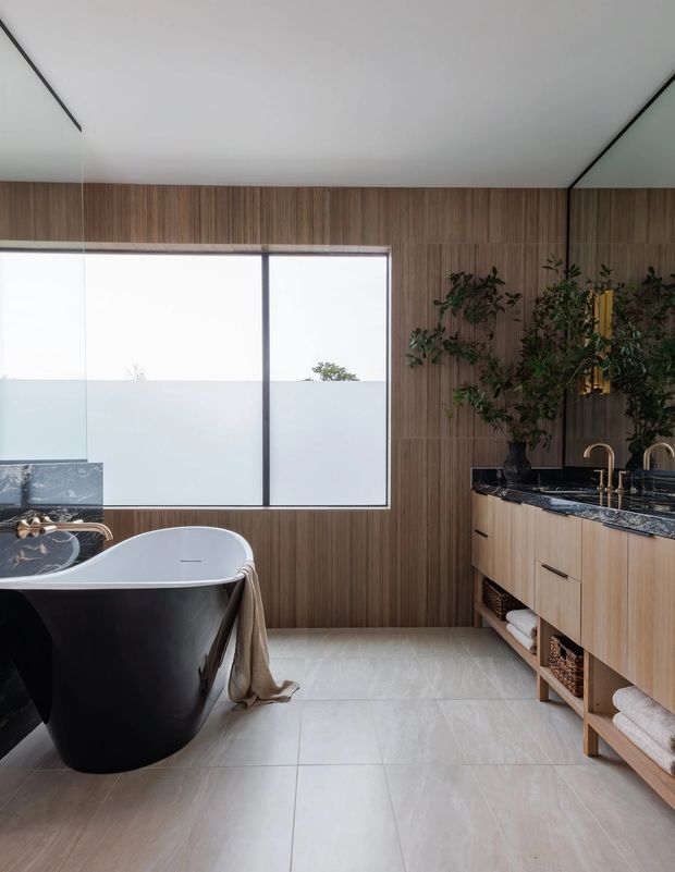 A refined modern bathroom featuring a freestanding black-and-white bathtub, a double vanity with light timber cabinetry and open towel storage, a dark stone benchtop, brass tapware, and extensive vertical timber wall panelling. Large frosted-glass windows provide soft natural light, while pale large-format floor tiles create a calm, contemporary atmosphere.