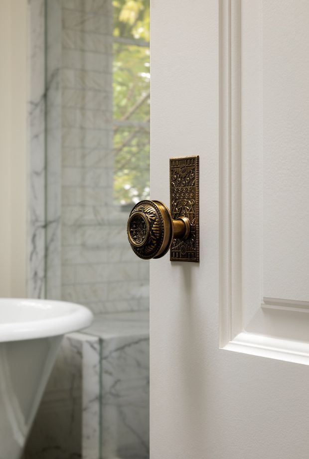 Close-up architectural interior detail of an ornate antique-style brass or bronze door knob and rectangular backplate on a white raised-panel door. Through the partially open door, a bathroom is visible with a white freestanding basin and pale veined marble wall and floor surfaces. Soft natural light enters from a window, creating a refined traditional atmosphere.