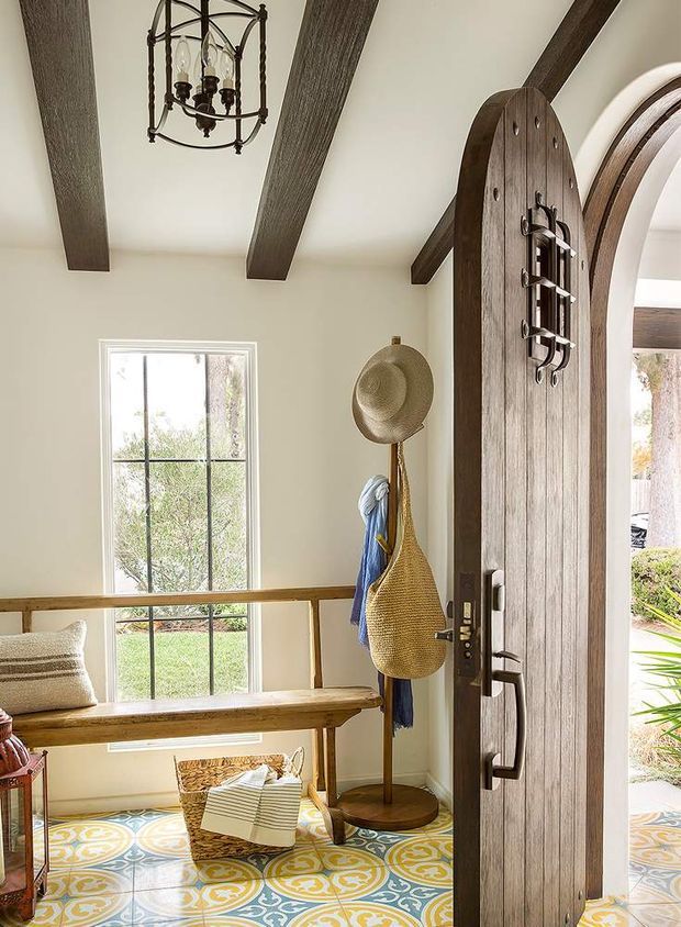 Bright residential entryway with a tall arched timber front door, dark exposed ceiling beams, cream walls, patterned encaustic-style floor tiles, and a built-in timber bench beneath a tall divided window. A vintage-style pendant light, coat stand, hats, woven bag, and small basket add a warm, rustic character. The open doorway reveals a small portion of landscaped exterior.