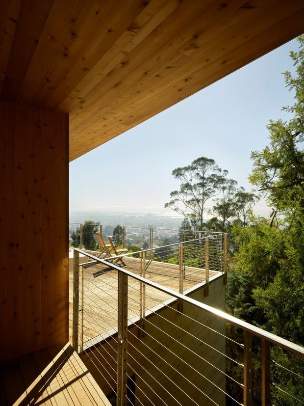 View from a timber-clad covered outdoor terrace or balcony toward a tree-filled, elevated landscape and distant urban vista. The space features warm timber decking and soffit, slim dark metal posts, and horizontal cable balustrades, with strong sunlight and a natural setting.
