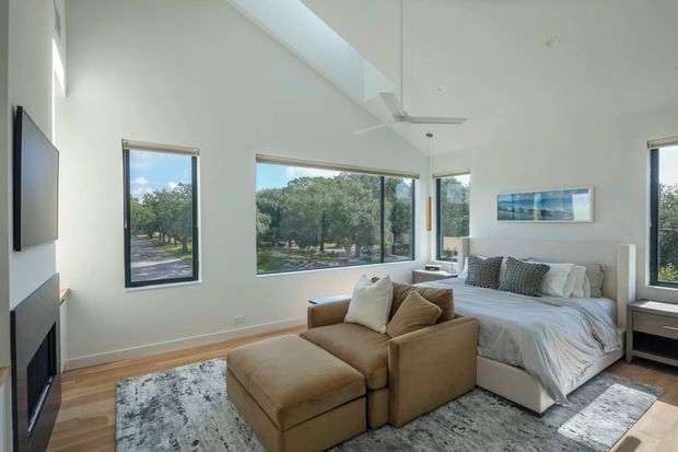 Bright contemporary bedroom with a high sloped ceiling, large windows overlooking trees, a centrally placed upholstered bed, and a tan modular lounge chair with ottoman in the foreground. The room features pale walls, timber flooring, grey patterned rugs, simple bedside furniture, recessed lighting, and a wall-mounted television with a fireplace below.