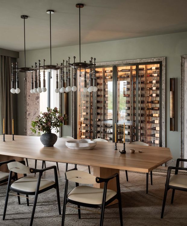 A refined dining room with a long light-wood dining table surrounded by sculptural black-framed cream chairs. Multiple linear pendant lights with globe bulbs hang above the table. The room features dark curtains, pale walls, textured stone-like wall panels, and a large illuminated glass-fronted wine storage display with extensive bottle shelving. Neutral layered finishes and warm lighting create a sophisticated, luxurious atmosphere.
