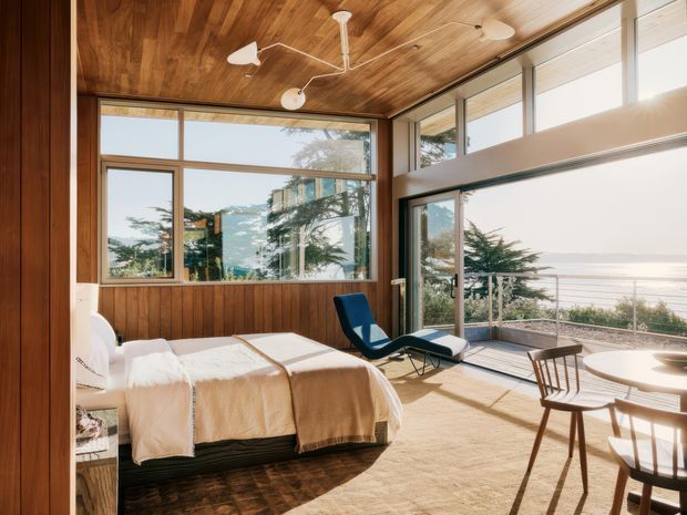 Sunlit coastal bedroom with floor-to-ceiling glazing overlooking the ocean. The room features warm timber wall and ceiling panelling, a large bed with neutral bedding, a deep blue lounge chair, and a compact timber dining or sitting area. Mid-century-inspired furniture and expansive windows create an airy, relaxed interior connected to the coastal landscape.