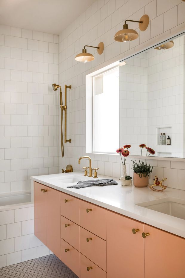 Bright bathroom featuring white rectangular wall tiles, a white countertop with integrated basins, peach-pink vanity cabinetry with brass hardware, brass wall sconces and shower fittings, a large mirror, and a frosted window. The floor uses small dark geometric tiles, with flowers and potted greenery adding minor decorative accents.
