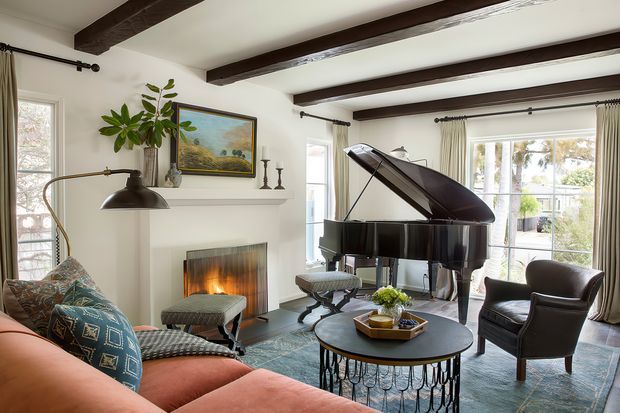 A bright, refined living room centred around a glossy black grand piano, with a fireplace, exposed dark timber ceiling beams, tall windows dressed in curtains, a patterned area rug, round coffee table, upholstered sofa and leather armchair. The room combines traditional architectural details with contemporary furnishings and abundant natural light.