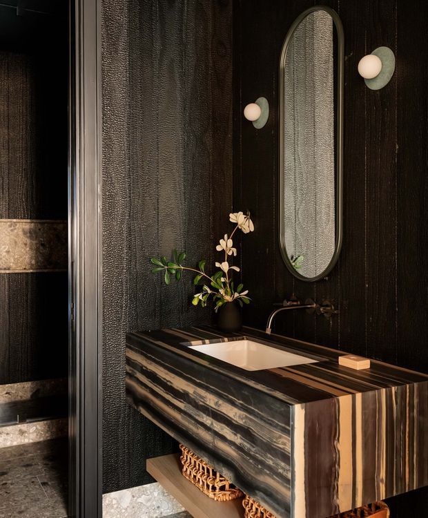 A moody, luxurious bathroom vanity with a dark textured wall finish, a sculptural striped timber or stone vanity, integrated white basin, bronze-toned wall-mounted tapware, oval mirror, and globe wall sconces. A partially visible shower area appears at left, with a small plant and woven baskets adding warmth.