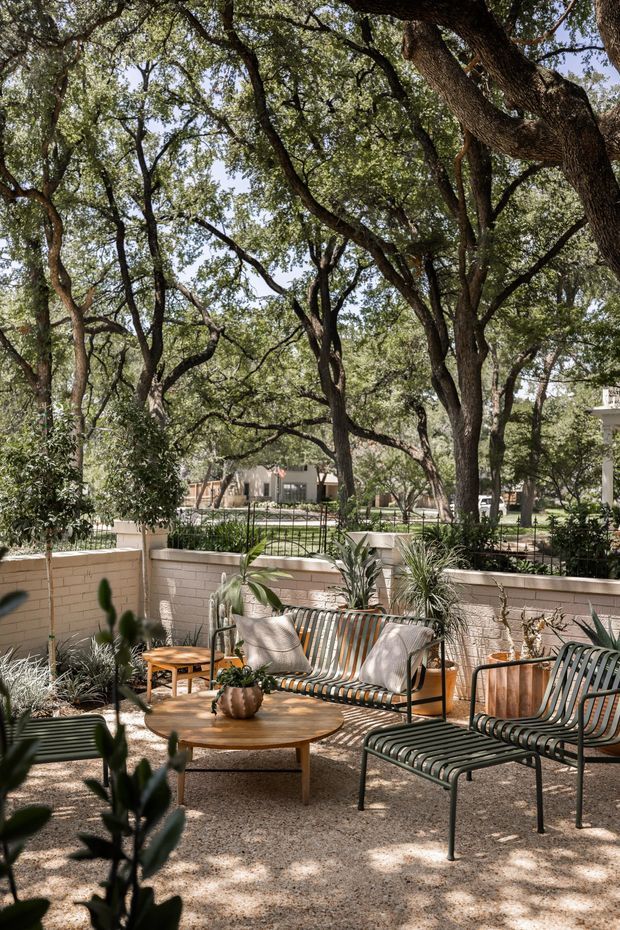 A shaded outdoor courtyard or patio furnished as a relaxed lounge, with a striped metal-framed sofa and matching lounge chairs arranged around a round wooden coffee table. Potted plants, gravel paving, a low pale brick wall, black metal fencing, and mature spreading trees create a leafy, dappled setting.