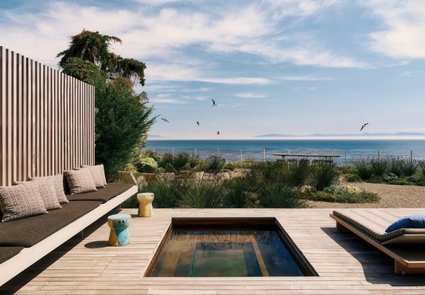 A coastal outdoor terrace overlooking the ocean, with a timber deck, built-in cushioned bench seating behind a vertical timber privacy screen, a small rectangular plunge pool or reflecting pool, and a low sun lounge. Native-style grasses and coastal planting separate the deck from the beach and water beyond. The scene has a relaxed, contemporary resort-like character with bright natural light and expansive sea views.
