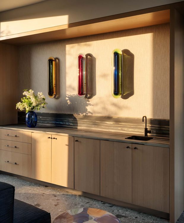 A warm, modern built-in kitchen or bar cabinetry wall with light timber drawers and cupboards, a dark stone benchtop and integrated backsplash, and a black undermount sink with tap. Three colourful sculptural wall lights or artworks are mounted above the counter, while sunlight creates strong shadows across the textured wall. A blue vase with flowers and partial seating are visible in the foreground.