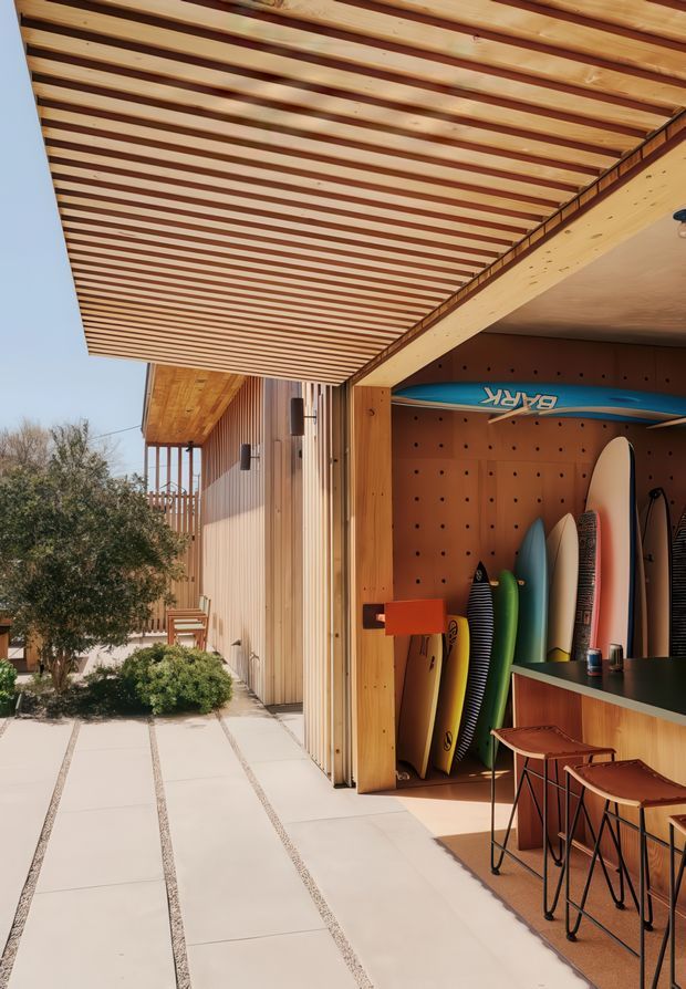 A warm, contemporary outdoor recreational space beside a timber-clad building. A deep slatted timber canopy projects over a pale paved walkway, leading to an open surfboard storage and casual bar area with a dark benchtop, timber cabinetry, and high stools. Multiple surfboards are displayed against a perforated timber backing, while landscaped planting and a timber privacy screen frame the side of the space. The design combines natural timber, clean linear detailing, and a relaxed coastal lifestyle aesthetic.