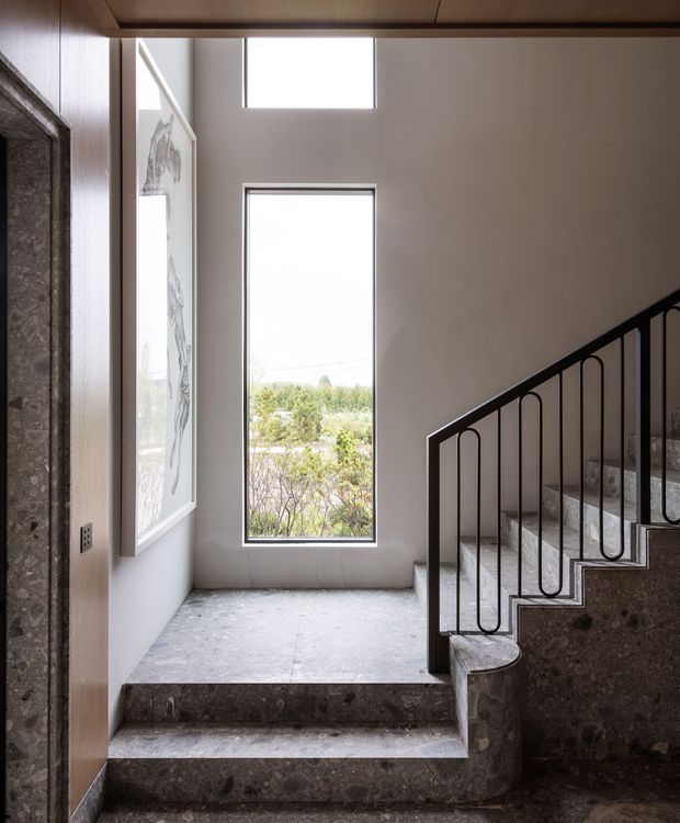 A refined interior stair landing with a short set of broad stone steps leading to a staircase. The space features dark textured stone flooring and stair risers, pale plaster walls, warm timber ceiling and wall lining, tall narrow windows, and a distinctive black metal balustrade with rounded vertical loops. A large framed artwork hangs on the left wall, while landscaped greenery is visible through the windows.
