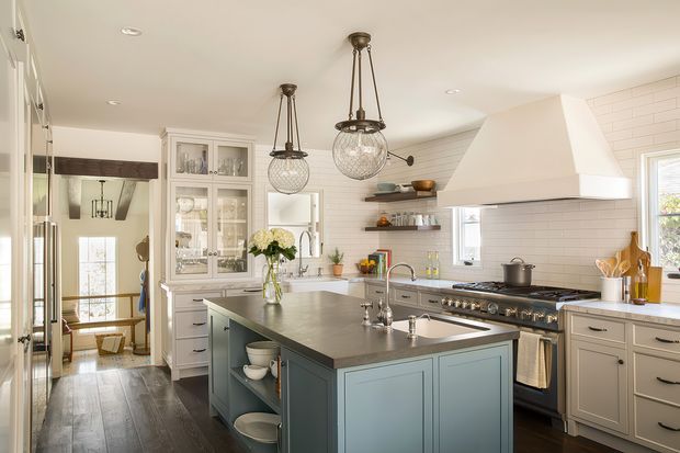 Bright, spacious traditional kitchen with a large muted blue-grey island, white shaker-style cabinetry, dark timber flooring, white horizontal wall tile, open shelving, and a large range cooker beneath a prominent custom rangehood. Two oversized glass pendant lights hang above the island, with a glazed display cabinet and adjoining hallway visible in the background.