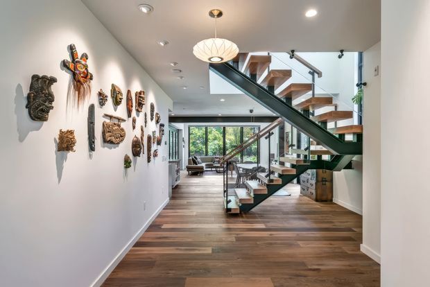 A bright, contemporary residential hallway and stairway with an open-riser floating staircase, dark green structural stringer and glass balustrades. Warm timber flooring continues through the space, while a white gallery wall displays a collection of decorative masks and carved artworks. The hall opens toward a living area with abundant glazing, and a sculptural pendant hangs above the circulation space.
