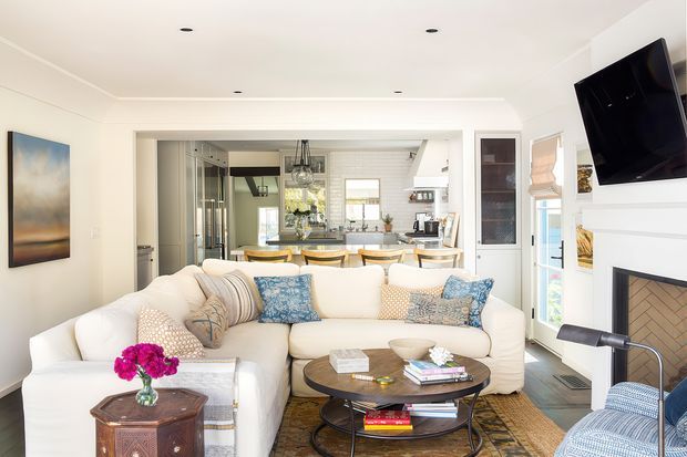 Bright, open-plan living room with a large cream sectional sofa, patterned cushions, round dark wood coffee table, accent chair, wall-mounted television and fireplace. The room opens directly toward a light-filled kitchen and dining area, with white walls, recessed lighting and large glazed doors/windows.