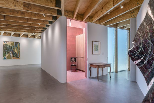 A spacious contemporary interior gallery or entry space with polished concrete flooring, white partition walls, exposed timber roof framing and plywood ceiling panels. The open-plan area includes colorful artwork, a narrow console table, a framed picture, and a view into a pink-toned adjoining room. A large abstract textile or artwork hangs on the right wall.
