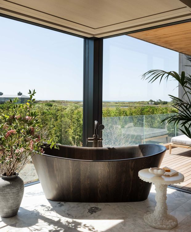 A luxurious freestanding bathtub sits in a bright bathroom beside expansive floor-to-ceiling glazing. The dark sculptural tub rests on a pale veined stone floor, with a small decorative side table and potted plant nearby. Black-framed glass doors open toward a timber deck and transparent balustrade, overlooking a broad coastal landscape. A timber-lined ceiling soffit and warm natural materials create a refined indoor-outdoor feel.