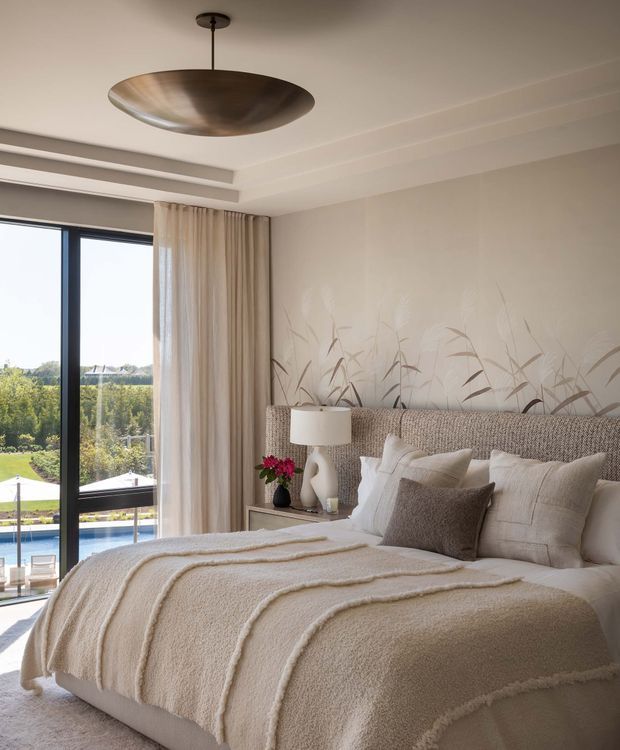 A bright, refined bedroom with a large upholstered woven headboard, layered cream and beige bedding, neutral bedside tables and lamps, and a delicate botanical wall mural. Full-height black-framed glazing and sheer curtains open to a leafy landscape and pool view. A sculptural bronze pendant light and soft cove ceiling detailing add warmth and polish.