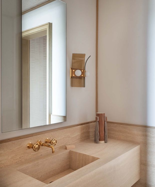Close-up of a refined bathroom vanity with a sculpted stone basin and matching stone countertop and backsplash. The space features warm beige travertine-like surfaces, a polished brass wall-mounted tap, timber trim, a large mirror, and a vintage-inspired wall light. The styling is restrained, warm, and luxurious.