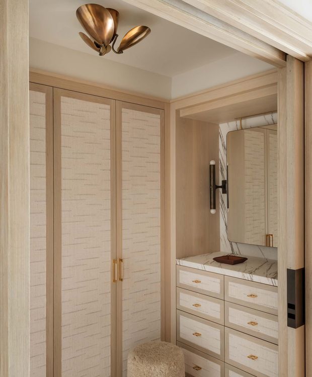 A refined dressing and vanity area viewed through a timber-trimmed doorway. Floor-to-ceiling light timber cabinetry with textured cream inset panels occupies the left side, while a built-in vanity with multiple drawers, a pale veined stone benchtop, rounded rectangular mirror, and wall light sits to the right. A sculptural bronze ceiling light and coordinated warm metal hardware add a luxurious accent. A small upholstered stool is positioned in front of the vanity.