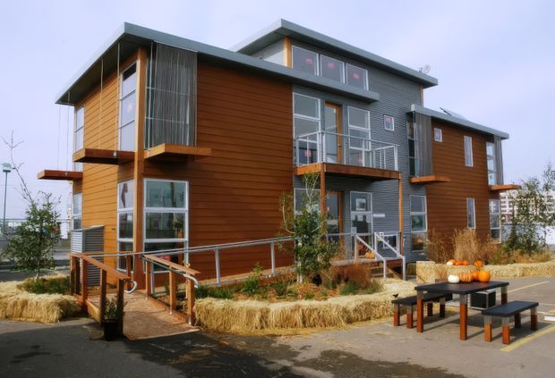 Exterior view of a contemporary multi-storey residential building with a stepped flat roof, warm horizontal timber cladding, dark grey metal cladding, extensive glazing, projecting balconies, and accessible ramped entry. The landscaped foreground includes raised planting beds and outdoor picnic-style seating.